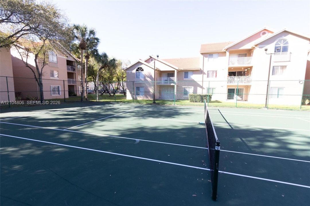 Hardcourt tennis court with net, surrounded by fencing and mature trees