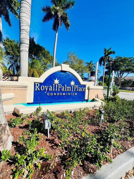 Monument sign featuring "Royal Palm Place Condominium" with blue and cream-colored finishes, set amidst lush landscaping and mature palm trees