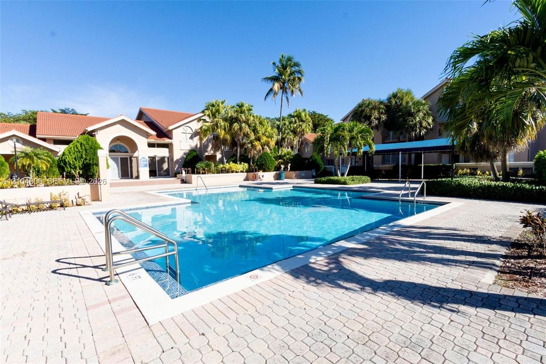 Resort-style swimming pool surrounded by paver patio and tropical landscaping, with community building featuring arched entryway