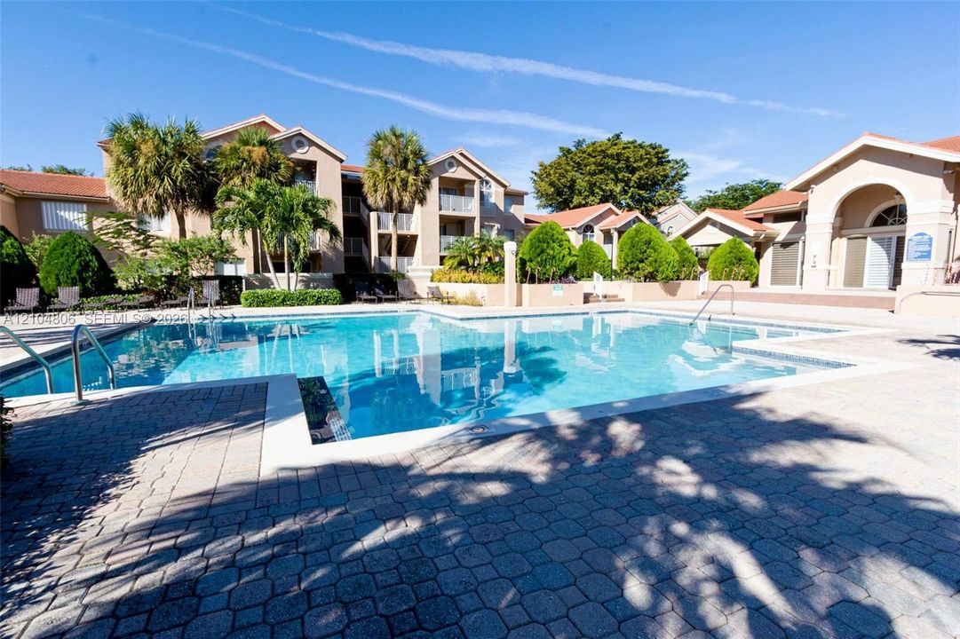 Expansive outdoor swimming pool with surrounding paver decking, adjacent to multi-story buildings with balconies, and palm trees