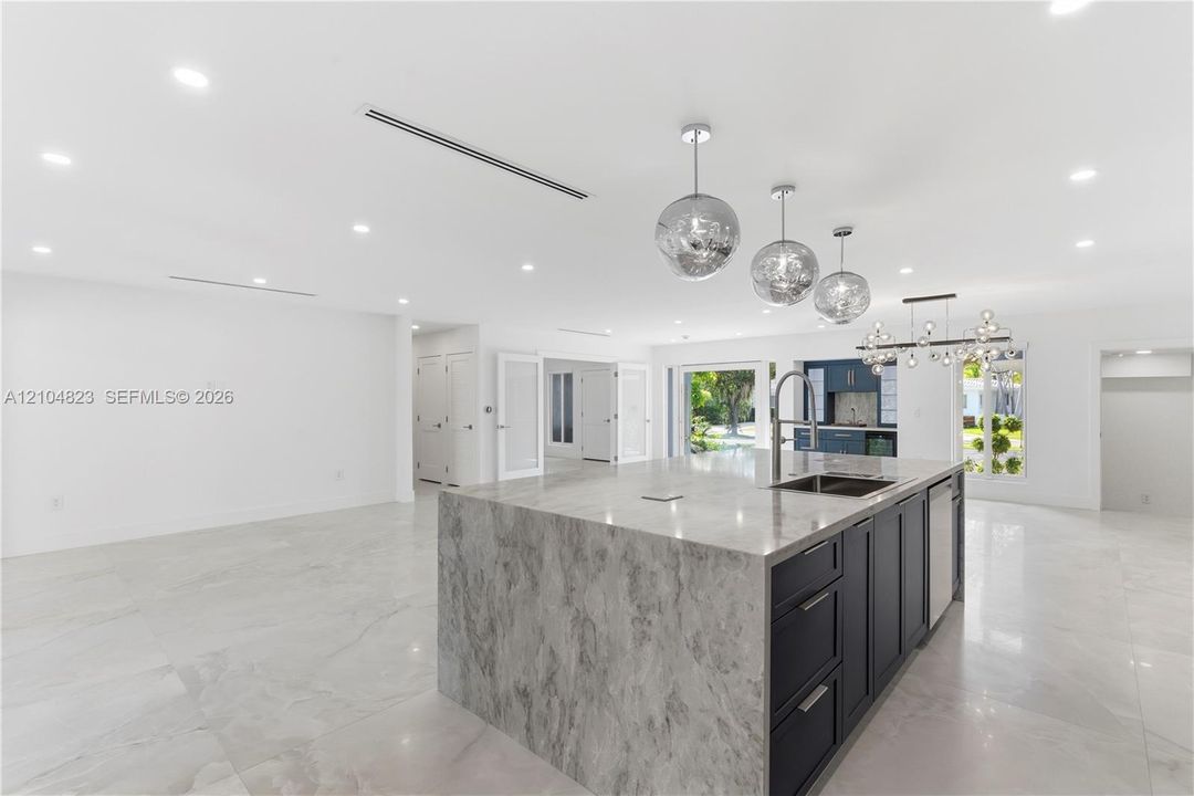 Kitchen featuring a waterfall-edge island with stone-finish countertop, dark-toned cabinetry, recessed lighting, metallic pendant fixtures, and large-format tile flooring