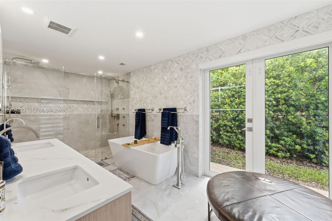Dual vanity with integrated sinks, freestanding soaking tub with floor-mounted faucet, glass-enclosed shower, wall-mounted towel bars, and French doors leading to exterior