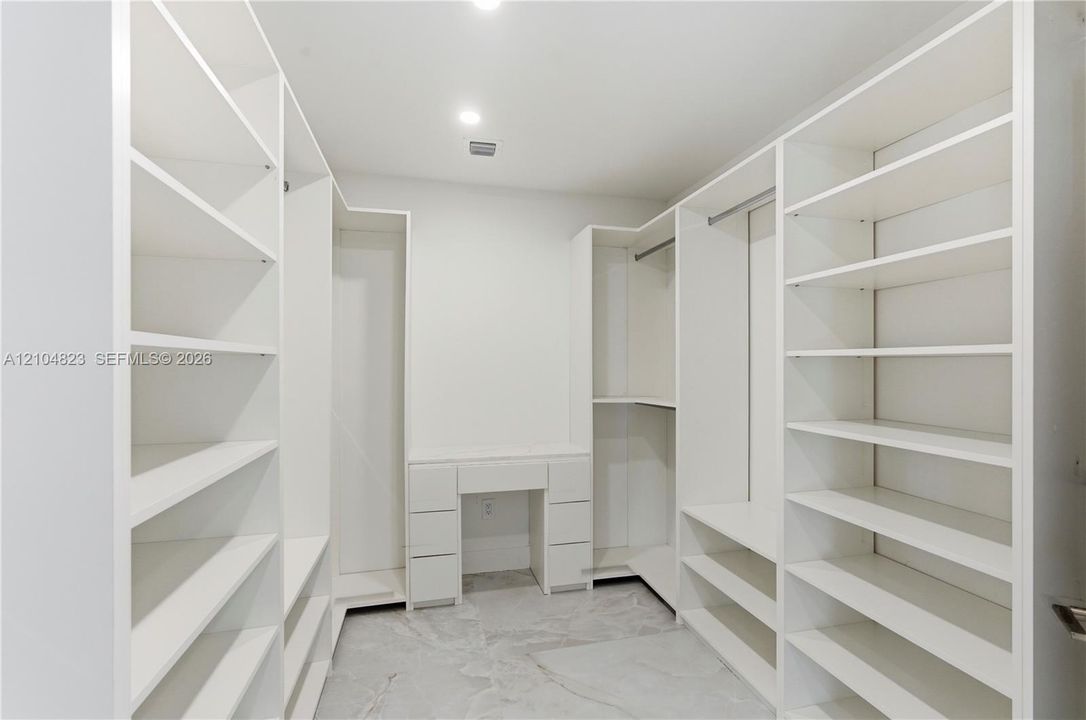 Walk-in closet featuring extensive built-in white shelving, integrated hanging rods, a central vanity with drawers, and polished stone-finish flooring