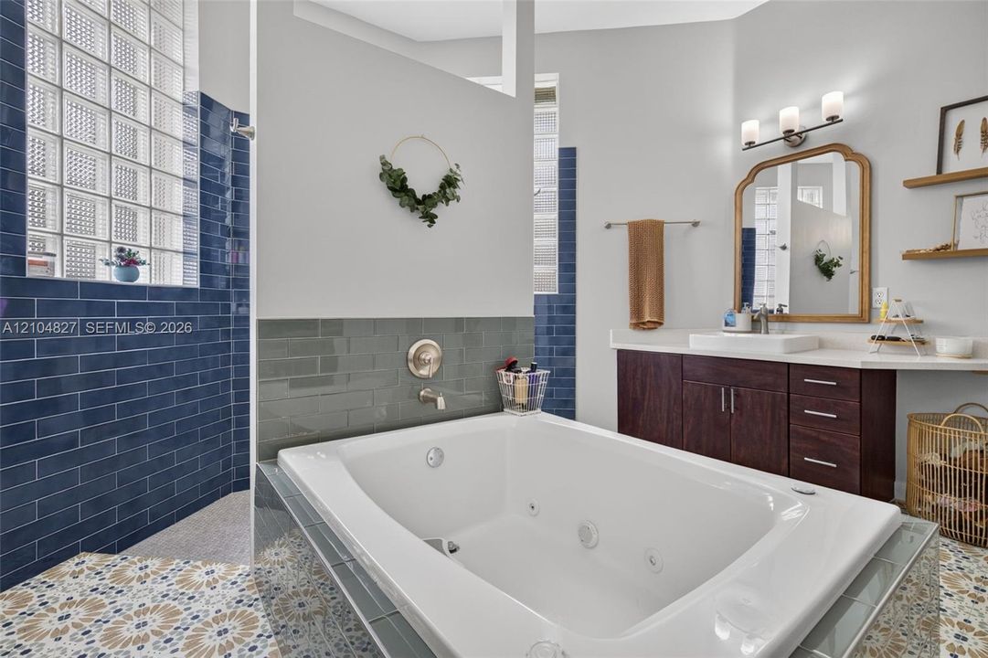 Jetted soaking tub featuring a gray tile surround, dark wood-finish vanity with a rectangular vessel sink, blue subway tile wall accents, glass block windows, and patterned floor tile