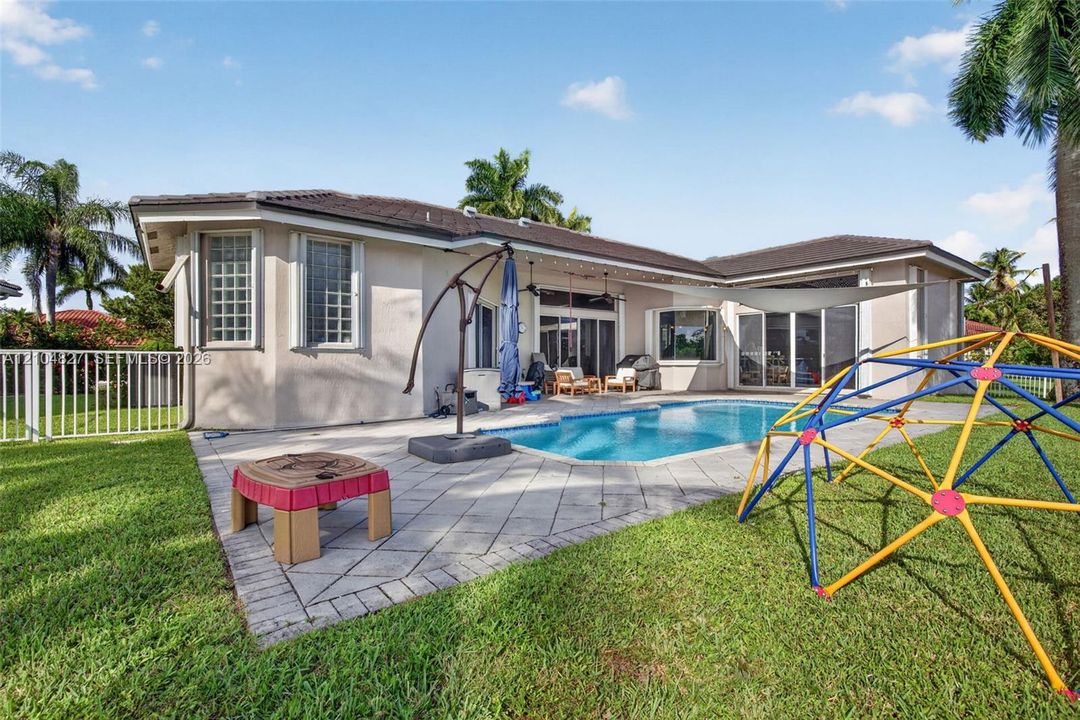 Rear exterior featuring a covered patio, paved stone deck, in-ground swimming pool, and white aluminum perimeter fencing