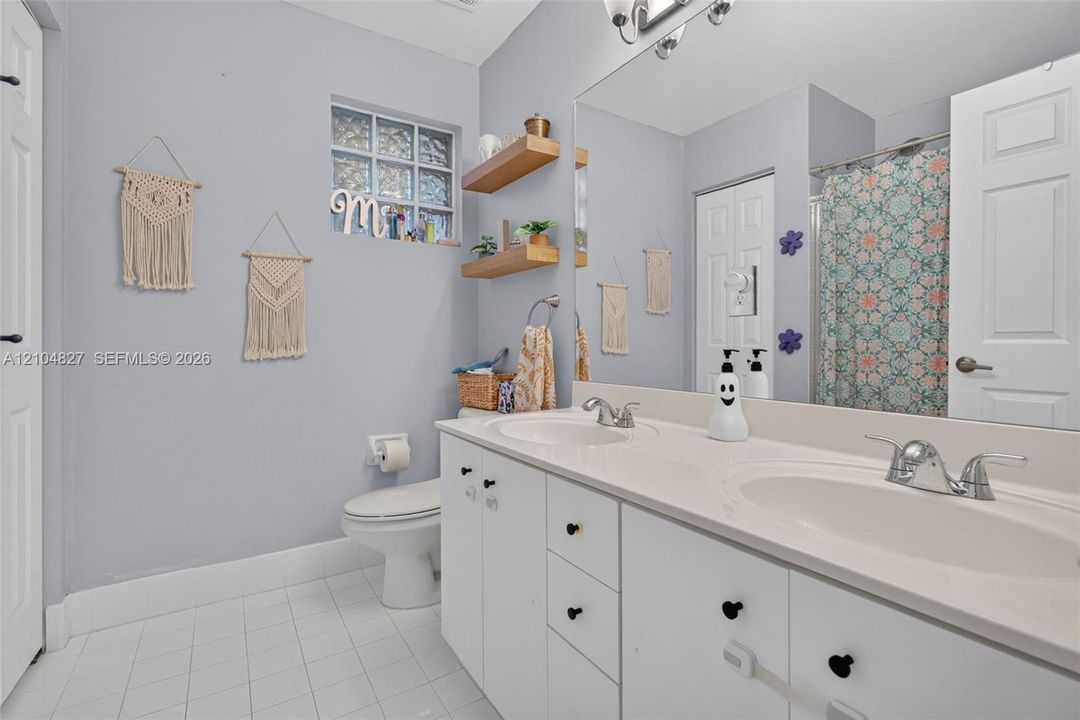 Bathroom featuring a dual-sink vanity with a solid-surface countertop, glass block window, wall-mounted wood shelving, and tiled flooring
