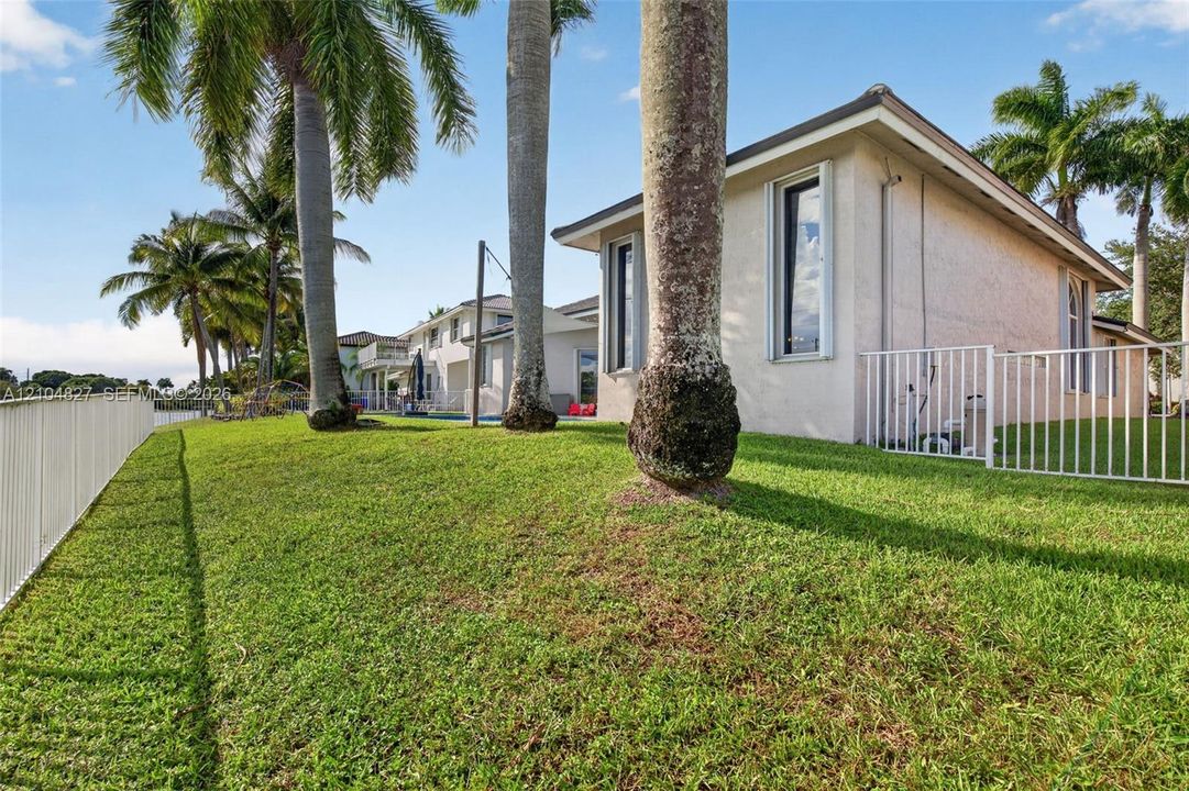 Stucco exterior residence featuring a sprawling lawn, mature palm trees, perimeter fencing, and water frontage