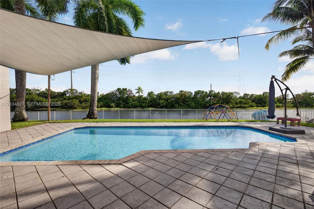 In-ground swimming pool with tile surround, expansive paver patio, installed shade sail, and white metal perimeter fencing overlooking the water