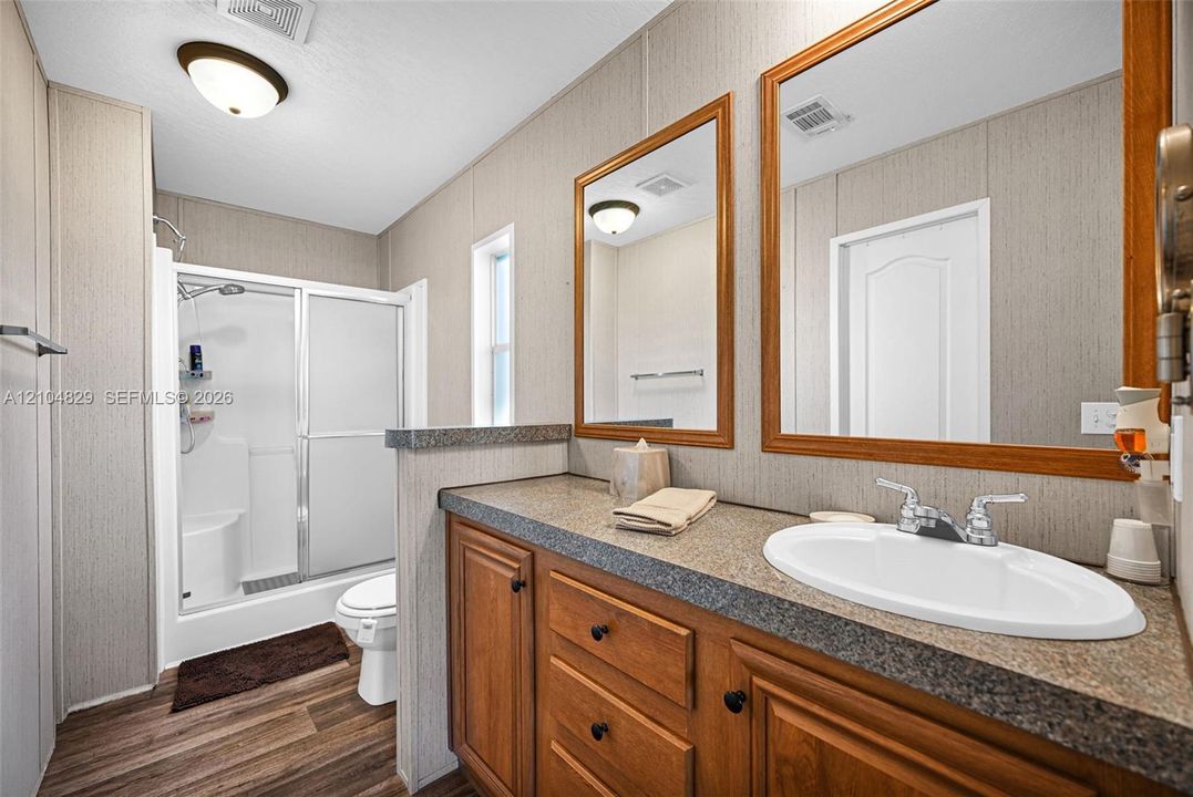 Bathroom featuring a wood-tone vanity with a dual-basin countertop, two framed mirrors, wood-finish flooring, and a walk-in shower enclosure