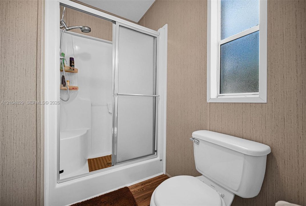 Walk-in shower enclosure featuring a built-in seat, adjustable shower head, and sliding glass doors, paired with a white porcelain toilet and vertical window with privacy glass