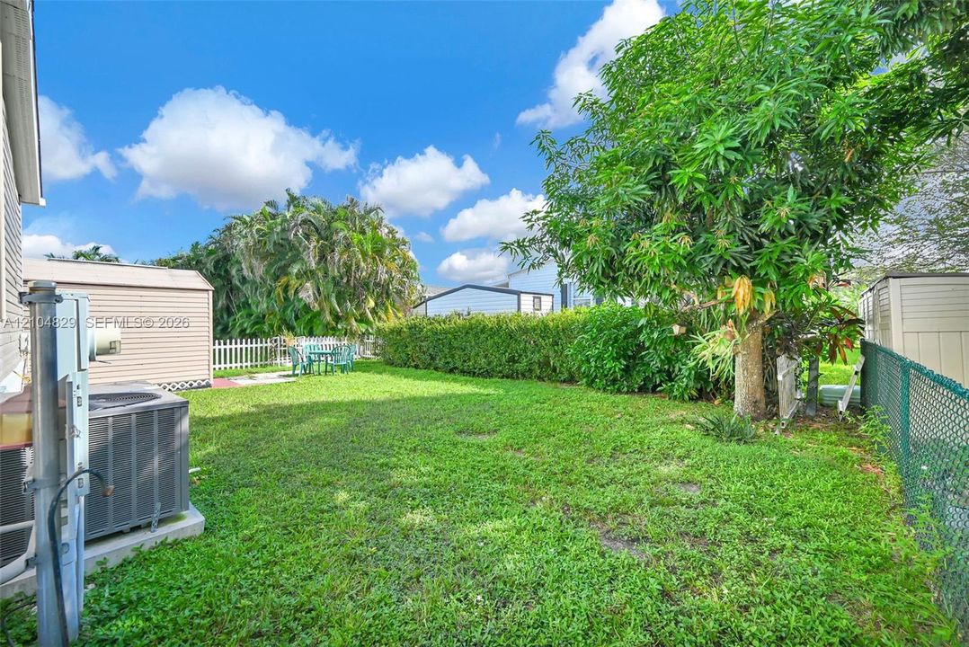 Expansive backyard with mature trees and lush lawn space, fenced perimeter, and adjacent residential structures visible in the distance under clear skies