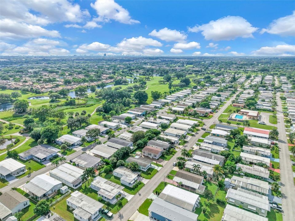 Aerial perspective featuring a residential community with paved streets, landscaped green spaces, established trees, and a central swimming pool