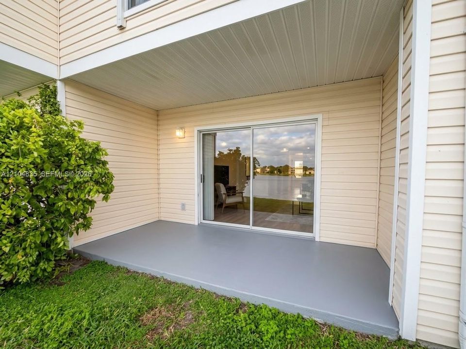Covered concrete patio with horizontal vinyl siding, sliding glass door, exterior wall-mounted light fixture, and water frontage