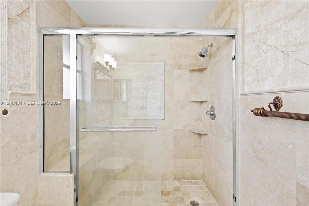 Walk-in shower featuring floor-to-ceiling stone tile, a glass enclosure with a polished metal frame, built-in stone shelving, and a wall-mounted shower head