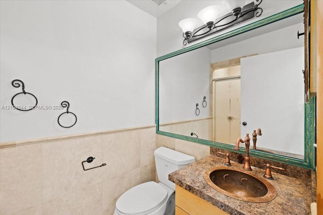 Bathroom vanity featuring a hammered copper drop-in sink, decorative stone countertop, wood-finish cabinetry, and a large wall-mounted mirror with a molded frame, complemented by half-height stone tile wainscoting and metallic wall-mounted hardware