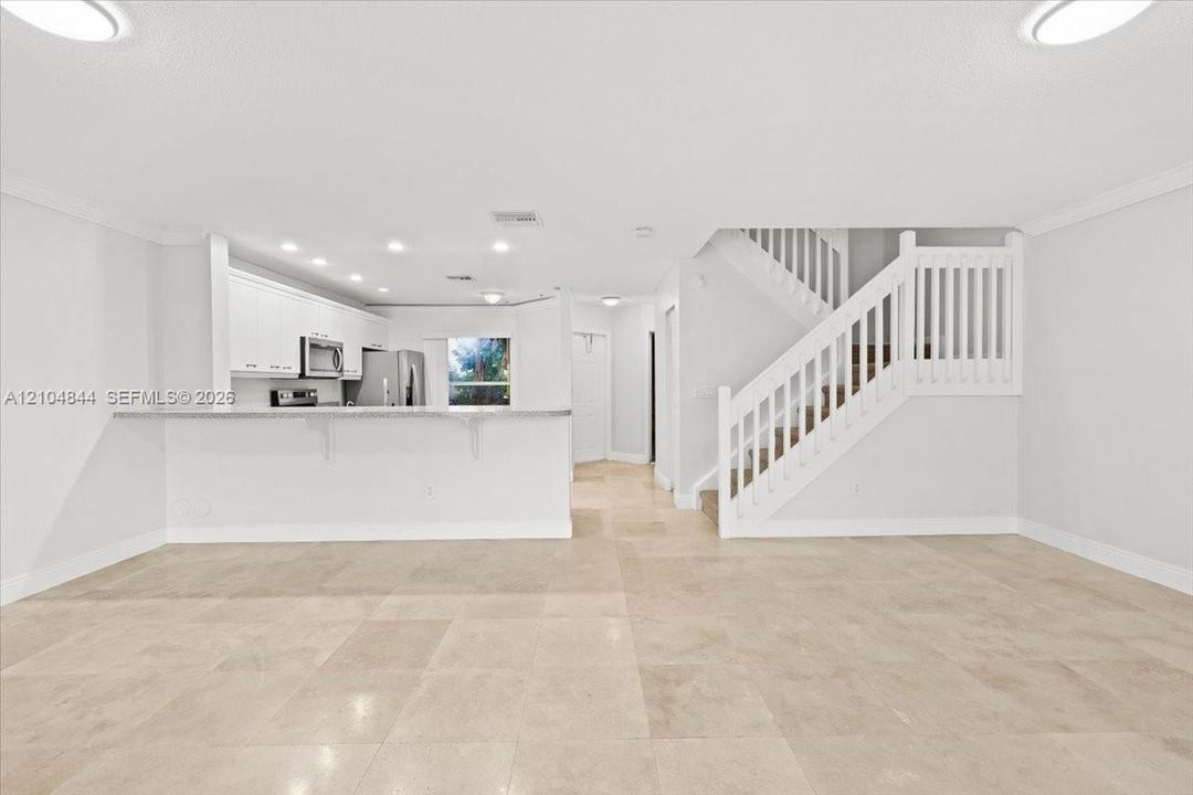 Open-concept interior featuring large-format tile flooring, white cabinetry, a breakfast bar with stone-finish countertops, recessed lighting, and a staircase with white balusters