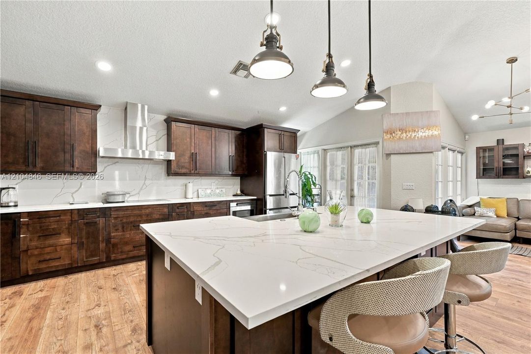 Kitchen featuring stained wood cabinetry, quartz countertops, a stone-patterned backsplash, stainless steel range hood, and pendant lighting over a central island