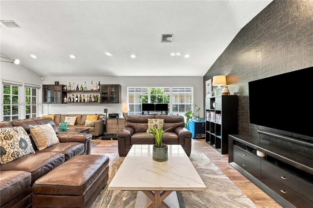 Vaulted ceilings with recessed lighting, integrated wall-mounted cabinetry, dark textured accent wall, wood-finish flooring, and expansive grid-pane windows