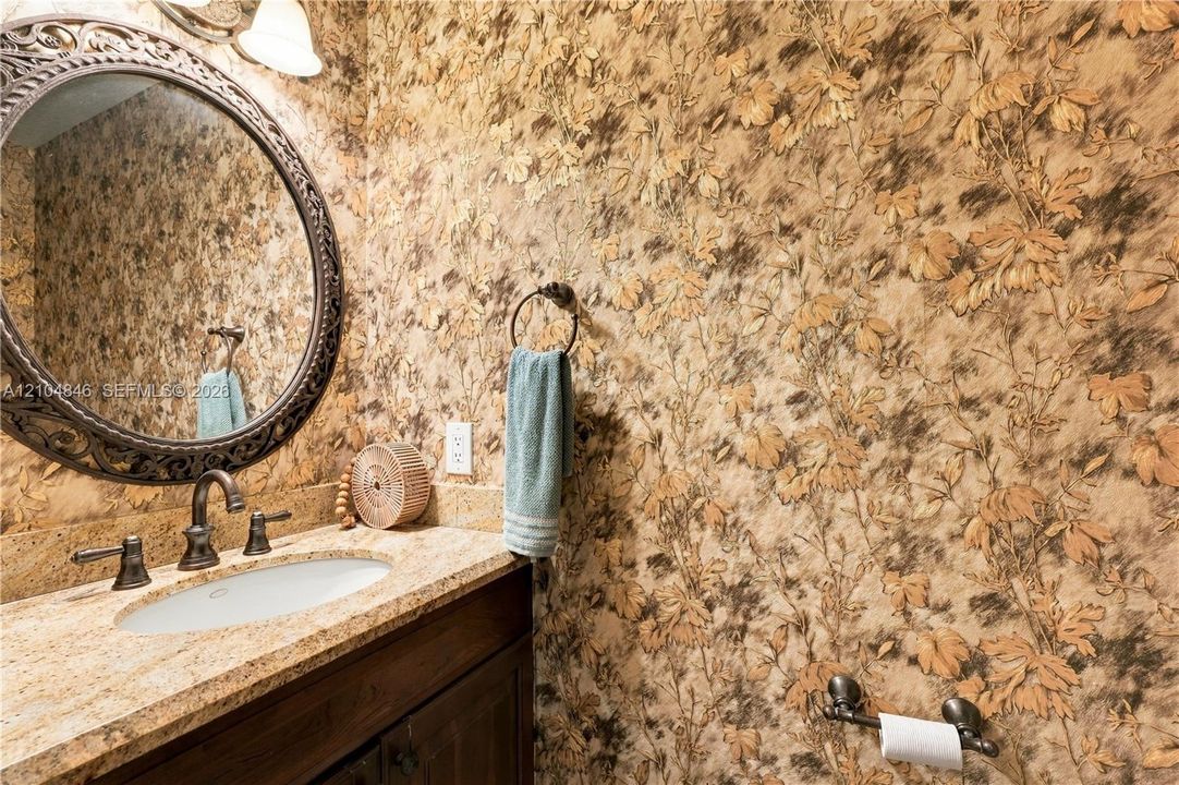 Half bath -Oval wall mirror with ornate metal frame, stone-finish vanity countertop, undermount sink, dark wood cabinetry, and patterned wallpaper
