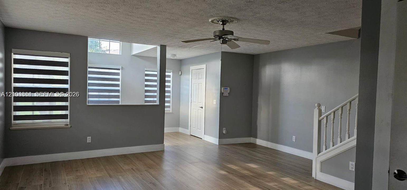 Wood-finish flooring, staircase with white balusters, ceiling fan with decorative medallion, multiple windows with blinds, and painted interior walls