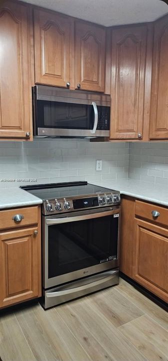 Stainless steel electric range, stainless steel microwave, light-toned wood-finish cabinetry, solid surface countertops, and light subway tile backsplash