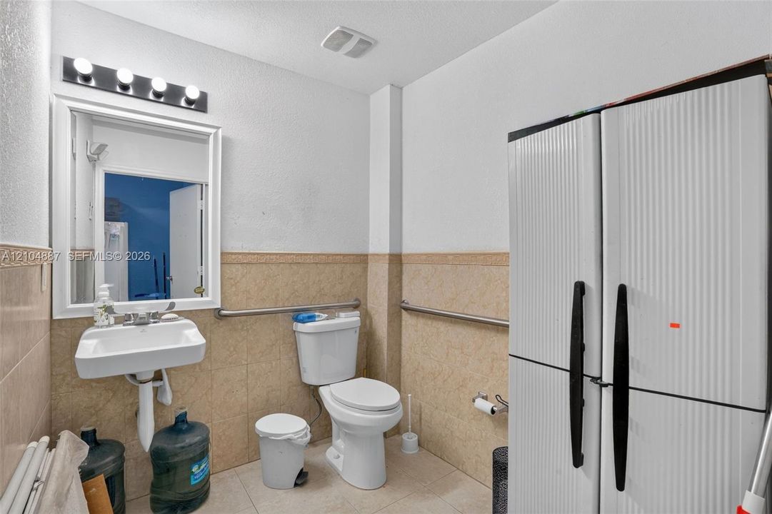 Wall-mounted sink with chrome fixtures, framed mirror with integrated four-bulb vanity lighting, ceramic tile wainscoting, and stainless steel grab bars