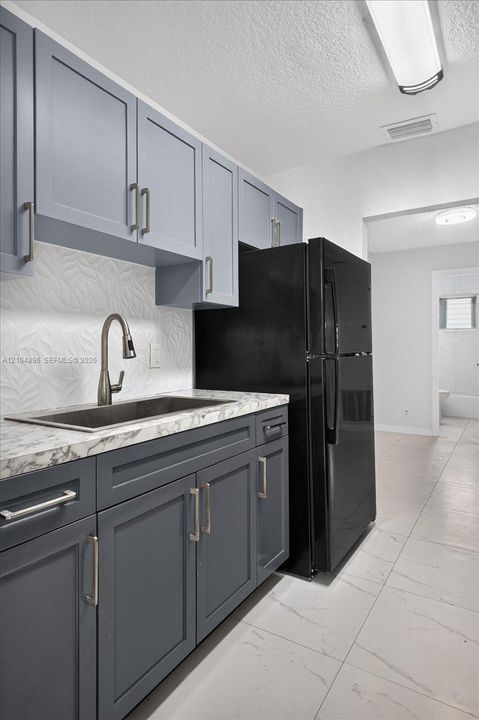 Kitchen featuring two-tone shaker-style cabinetry, stone-patterned countertops, a textured tile backsplash, an undermount sink with a gooseneck faucet, and large-format tile flooring