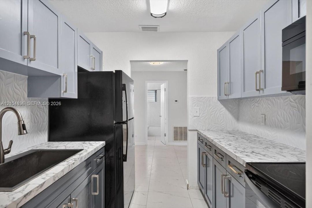 Kitchen featuring light blue shaker-style cabinetry, stone-patterned countertops, textured tile backsplash, dark-finished hardware, and ceramic tile flooring