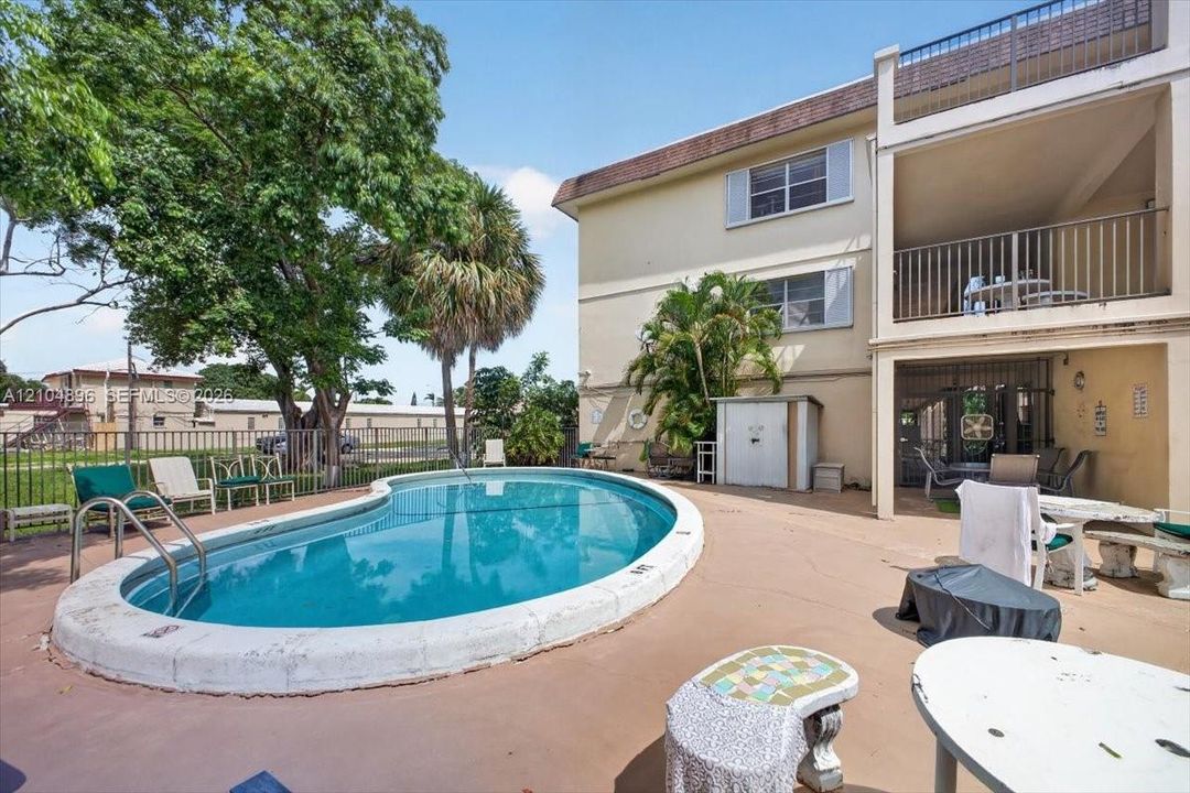 Kidney-shaped in-ground swimming pool with metal ladder, expansive concrete pool deck, multi-level exterior balconies with metal railings, and mature palm trees