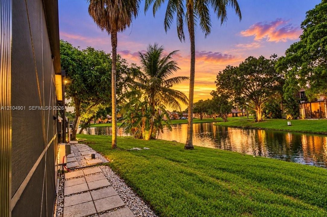 Waterfront backyard featuring a manicured lawn, palm trees, stone-paver walkway, and stone-aggregate ground cover