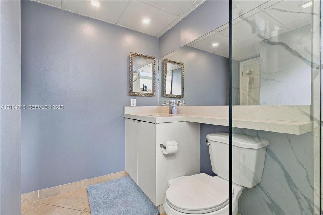 Built-in vanity featuring a light-toned countertop, large wall-mounted mirror, glass-enclosed shower with stone-finish surround, and tile flooring
