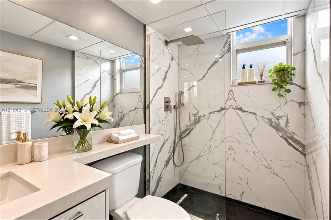 Bathroom featuring a walk-in shower with large-format stone-pattern tile, a frameless glass enclosure, a polished metal rainfall showerhead, and a vanity with a solid-surface countertop and full-width mirror