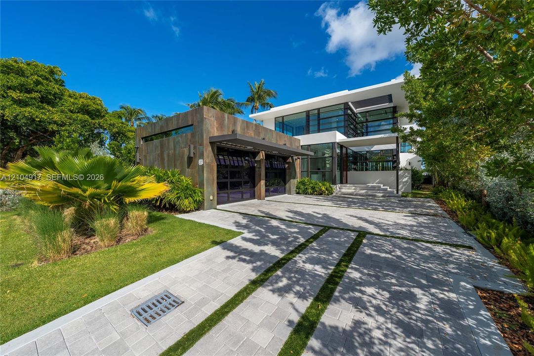 Modern architectural residence featuring metallic-finish garage cladding, expansive floor-to-ceiling glass paneling, multi-level structural design, and a paved stone driveway with turf accents