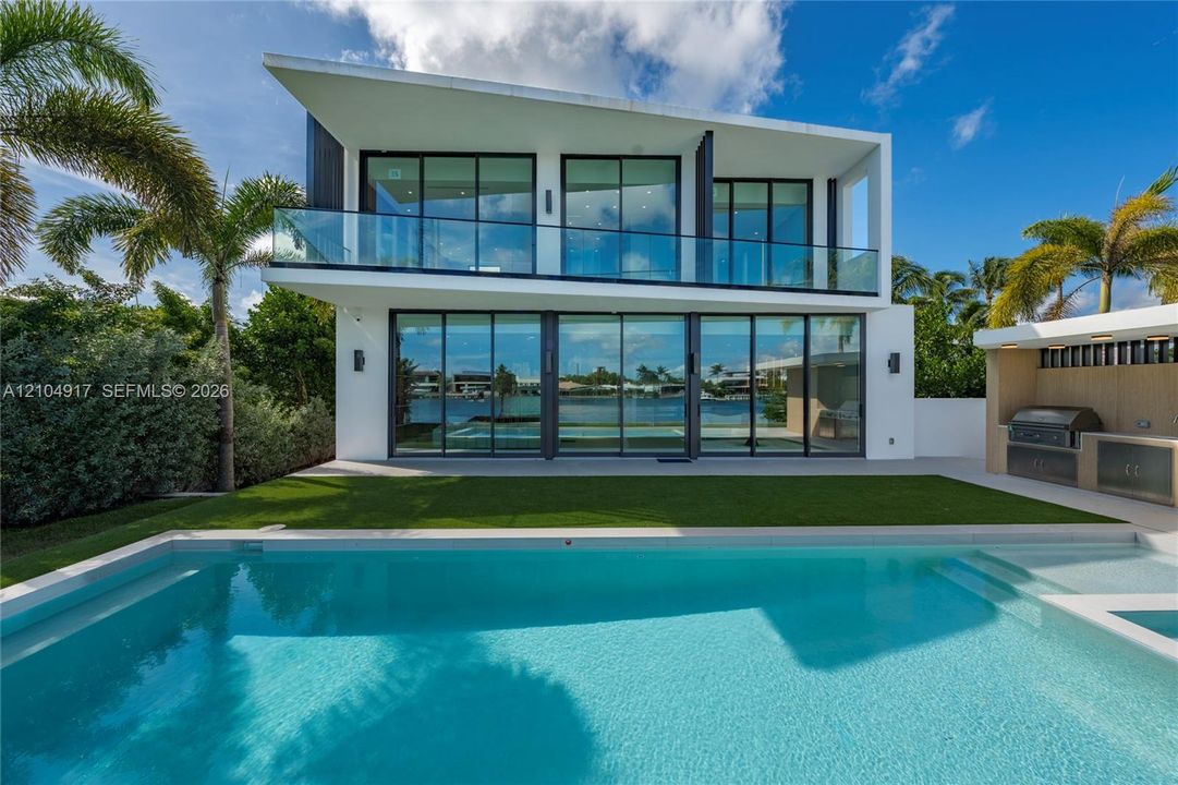 Modern two-story architectural exterior featuring floor-to-ceiling glass panel walls, a cantilevered balcony with a glass railing, a private in-ground swimming pool, and an outdoor kitchen area with a built-in grill