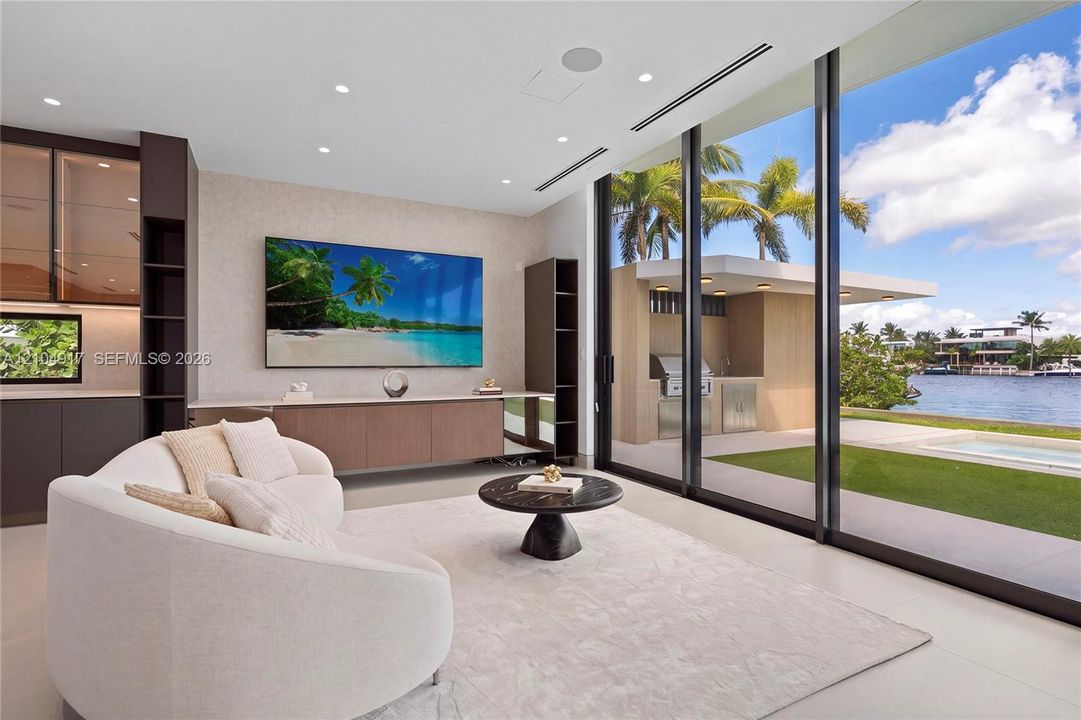 Floor-to-ceiling sliding glass doors, built-in media console, recessed lighting, integrated shelving, and an outdoor kitchen with water frontage
