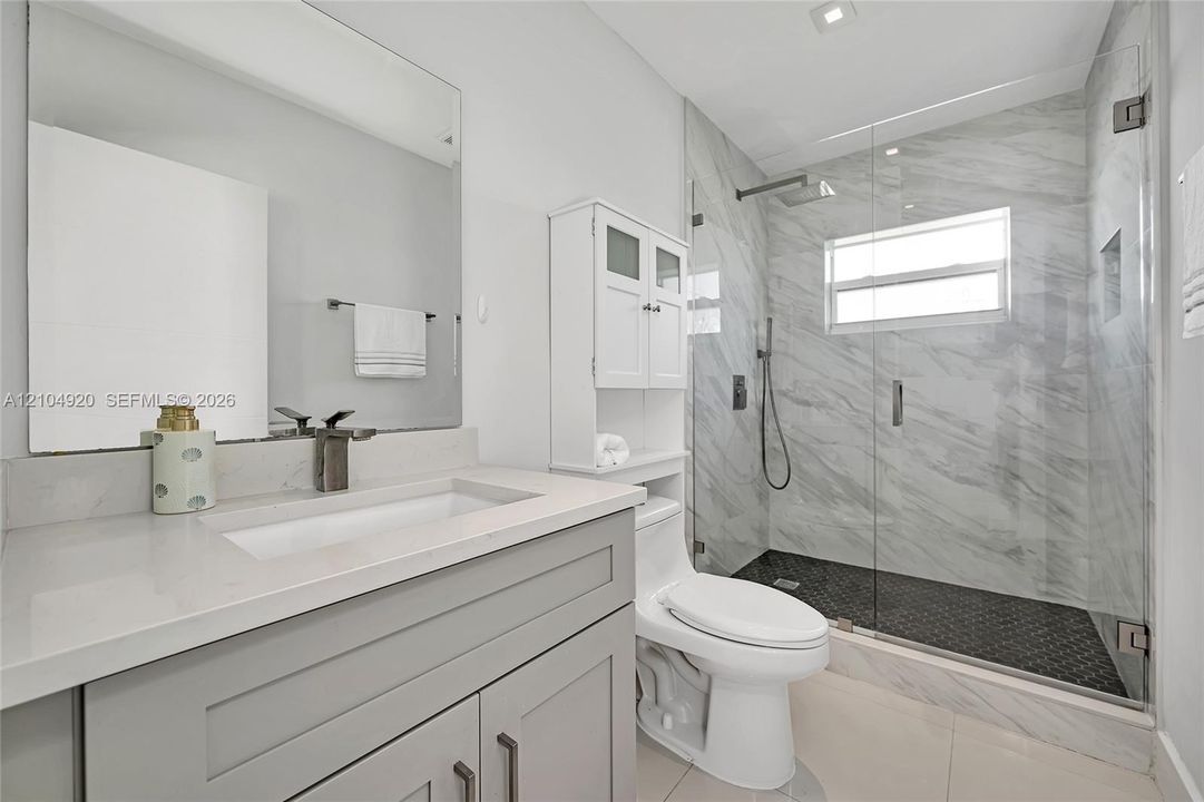 Bathroom featuring a grey vanity with a rectangular sink, stone-finish countertop, walk-in shower with floor-to-ceiling tile, and a wall-mounted glass enclosure