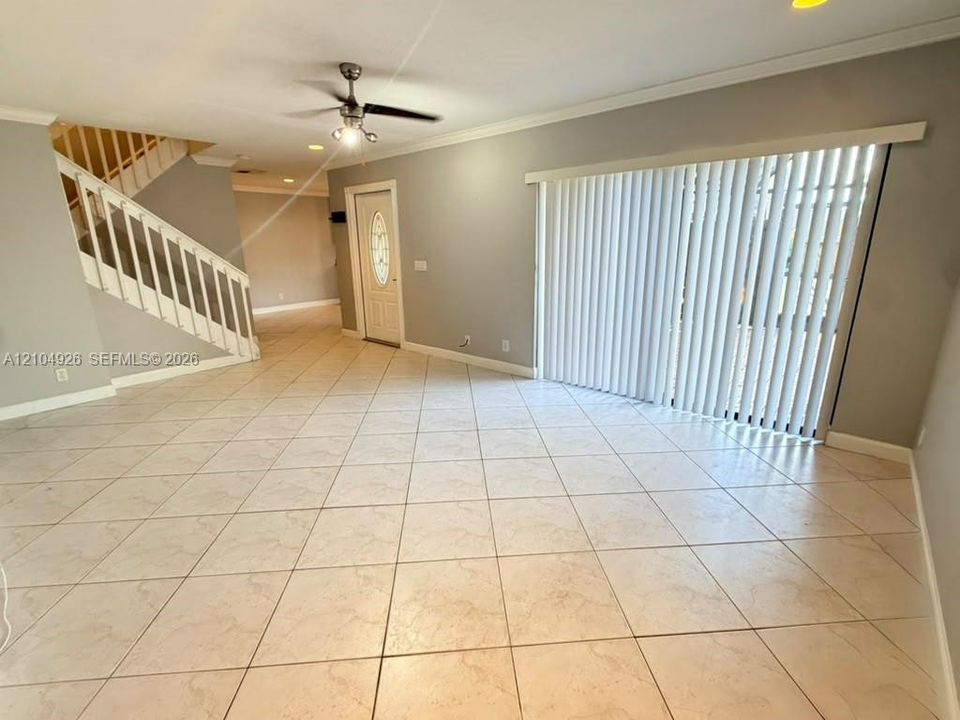Open-concept interior featuring large-format ceramic tile flooring, staircase with white railing, sliding glass doors with vertical blinds, and a ceiling fan