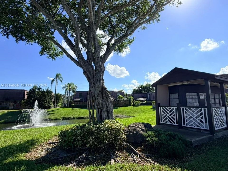 Community grounds featuring a water fountain, mature tree, manicured lawn, and covered mail kiosk with decorative lattice railing