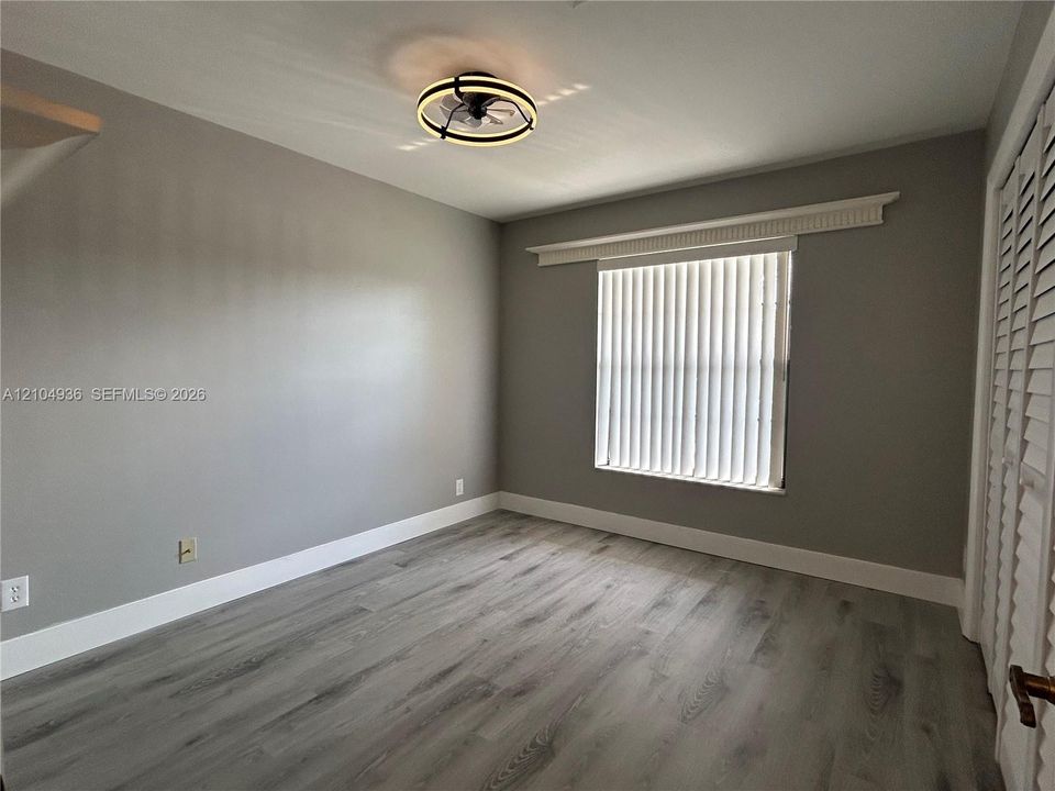 Room featuring wood-finish flooring, neutral wall paint, a circular flush-mount ceiling light with an integrated fan, a window with vertical blinds, and louvered bifold closet doors