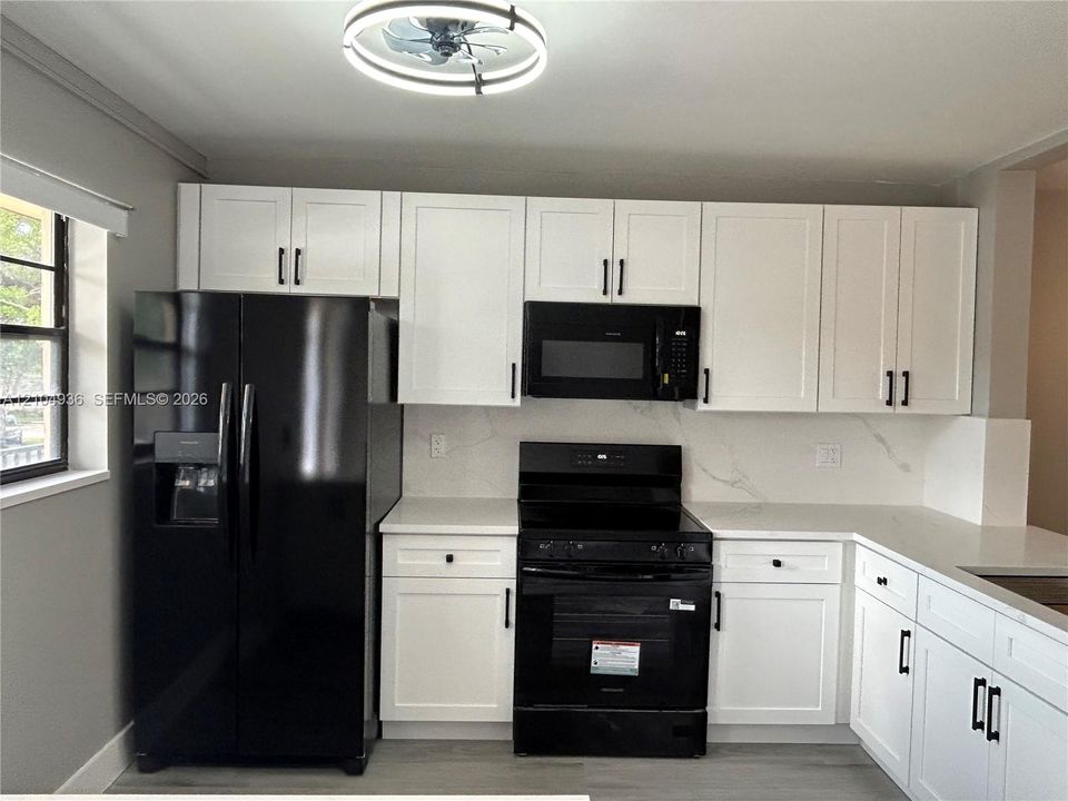 Kitchen featuring white shaker-style cabinetry, black hardware, solid-surface countertops with backsplash, black built-in appliances, and a circular LED ceiling fixture