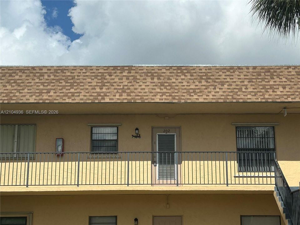 Exterior facade featuring a shingled roof, stucco wall finish, metal balcony railing, and windows with security bars