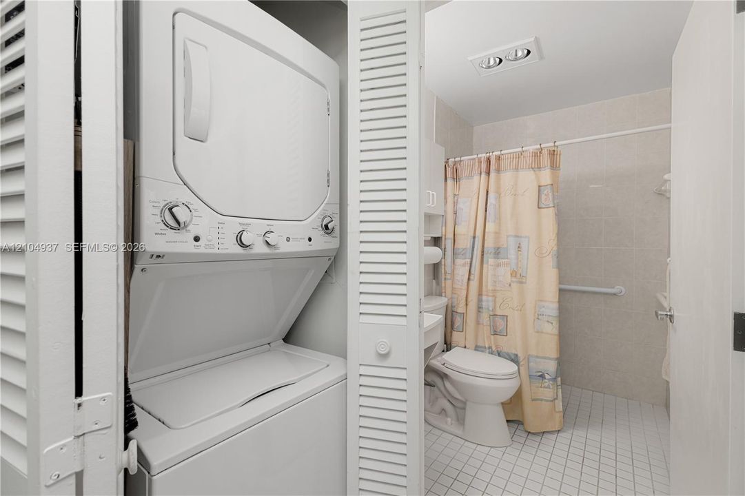 Stacked laundry unit housed in a louvered closet, bathroom featuring a tiled shower enclosure, white porcelain toilet, and grid-patterned floor tile