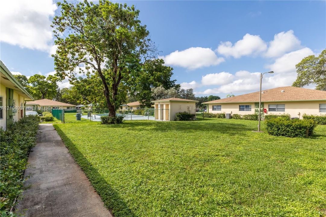 Expansive common area featuring a manicured lawn, concrete walkways, mature shade trees, a swimming pool, and an accessory storage structure