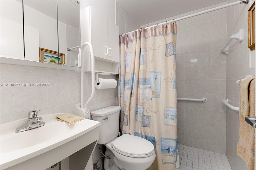 Bathroom featuring floor-to-ceiling tile, white vanity with an integrated basin, mirrored medicine cabinet, built-in upper wall storage, and wall-mounted support bars