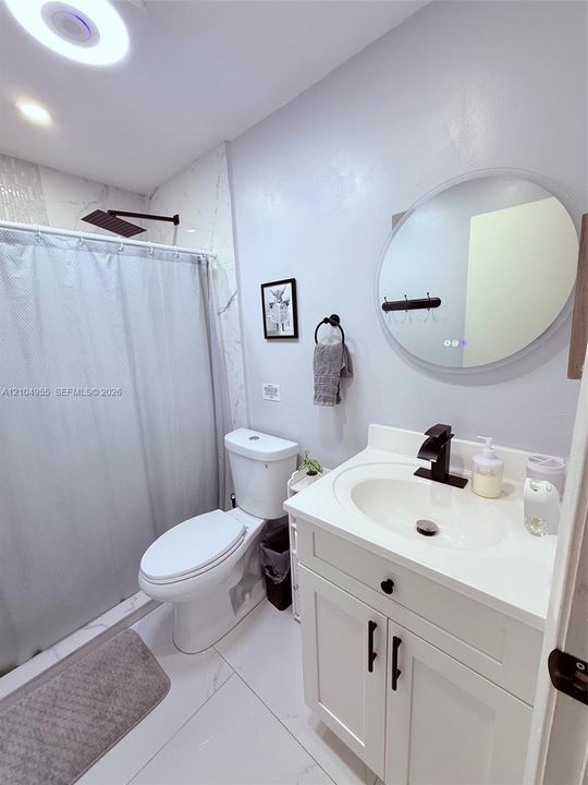 Bathroom featuring a white shaker-style vanity with an integrated basin, matte black faucet, wall-mounted round backlit mirror, and a walk-in shower with large-format stone-patterned tile and a rainfall showerhead