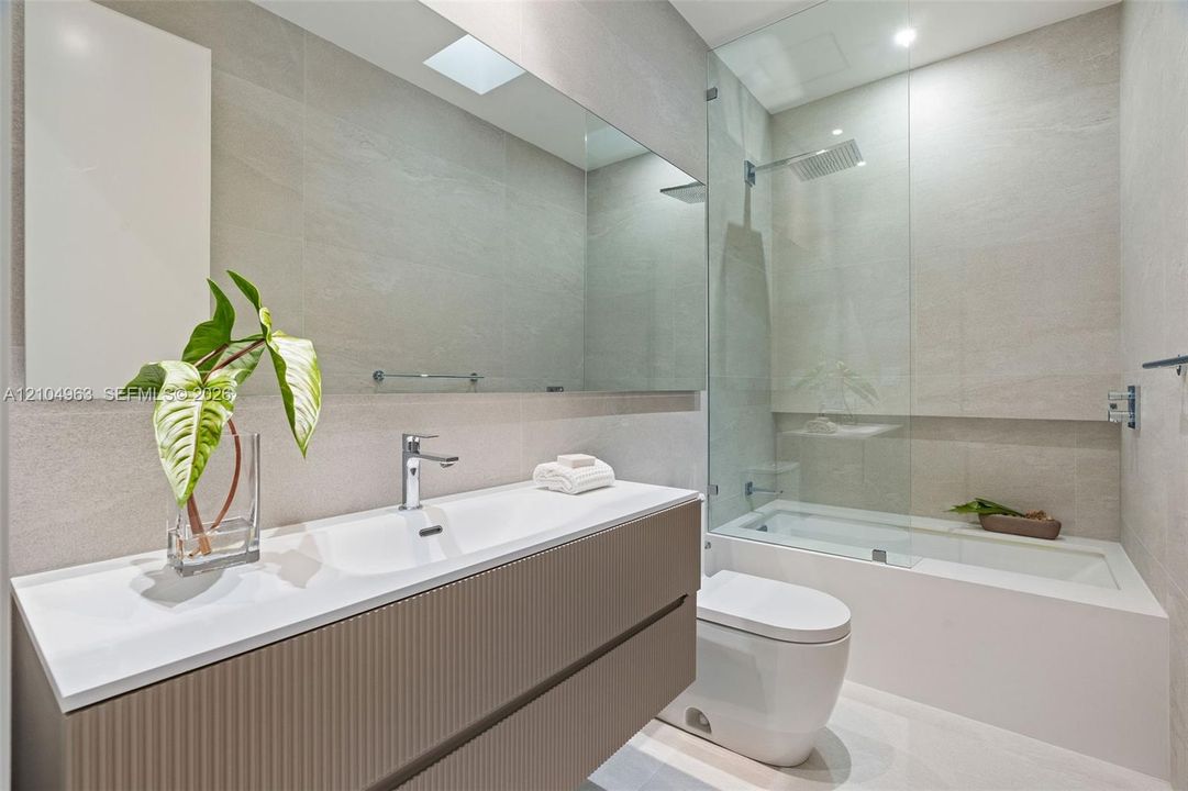 Floating vanity featuring a ribbed textured finish, integrated white basin, wall-to-wall mirror, glass-enclosed shower and tub combination, and full-height neutral wall tile