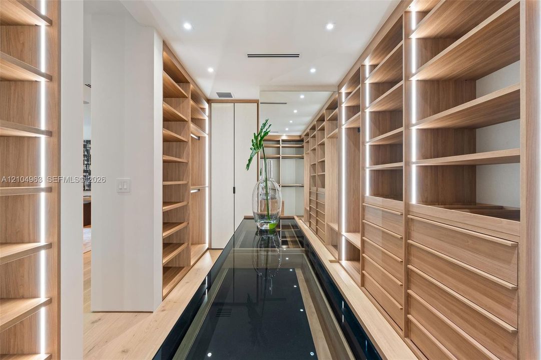Custom storage cabinetry featuring light wood-finish shelving, integrated vertical LED lighting, a glass-topped center island, and recessed ceiling lighting