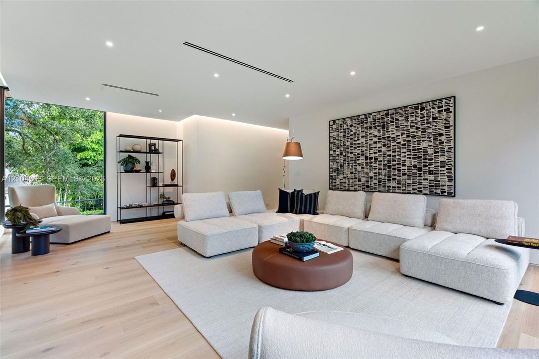 Living area featuring light wood-finish flooring, recessed linear lighting, hidden cove accent lighting, and a large floor-to-ceiling glass panel