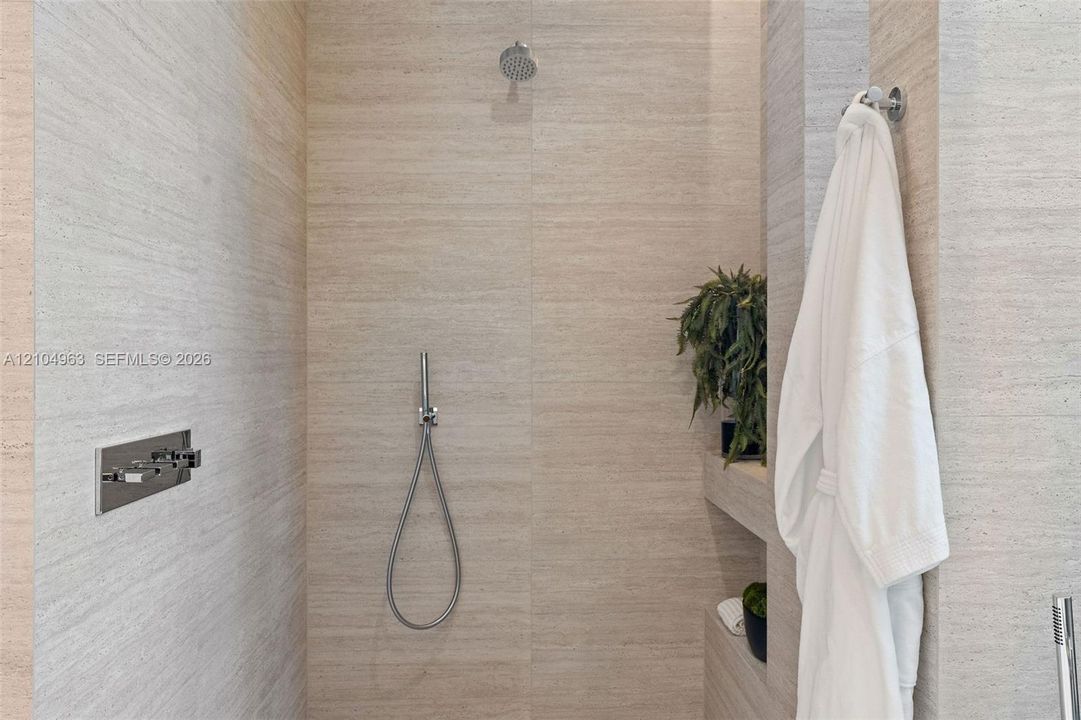 Walk-in shower featuring large-format textured stone wall tile, built-in shelving, a rainfall showerhead, and a wall-mounted handheld shower fixture with chrome hardware