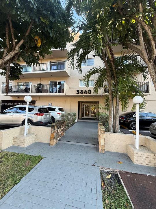 Multi-story building facade featuring private balconies, masonry retaining walls, a paved walkway, and mature palms